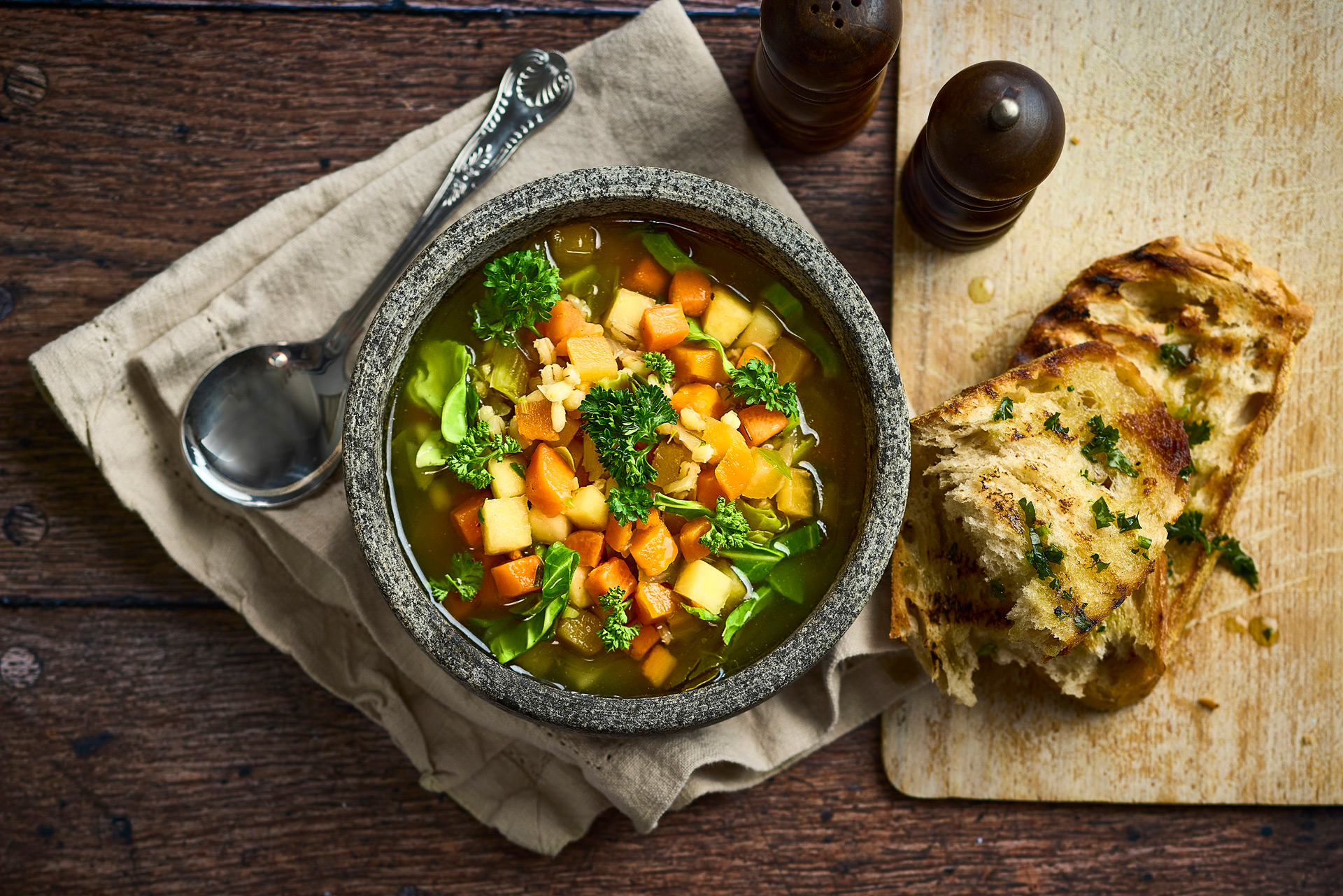 A bowl of vegetable soup on a table, with spoon, bread, and condiments